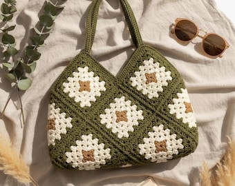 May include: A handmade green crochet tote bag with a floral pattern in white and brown. The bag has a sturdy handle and is displayed on a neutral-colored fabric. Sunglasses and a book are also in the image.
