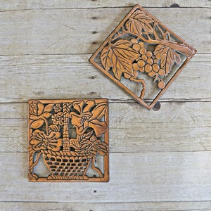 Könnte beinhalten: Zwei dekorative, quadratische Wandplatten aus Holz mit warmbraunem Finish. Eine Platte zeigt einen Blumenkorb, die andere eine Traube und Blätter. Die Platten sind vor einem hellen Holzhintergrund platziert.
