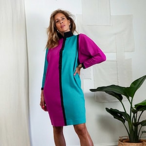 May include: A color-blocked long-sleeve dress featuring a vertical split design with one side in magenta and the other in teal. The dress includes a black mock neck collar, a central black button placket, and black trim on the sleeve cuffs.