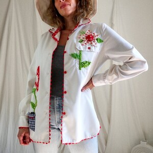 Peut inclure: Une chemise boutonnée blanche avec des fleurs brodées rouges et vertes. La chemise a une bordure en pompons rouges et une petite poche en vichy bleu et blanc.