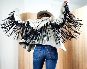 Pode incluir: Uma jaqueta de couro branca com franjas pretas e bordados florais. A jaqueta é usada sobre calças jeans azuis de perna larga.