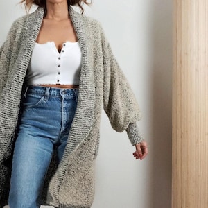 May include: A long, open-front cardigan in a speckled gray and beige knit. The cardigan has wide sleeves and a relaxed fit. Worn with a white crop top and blue jeans.