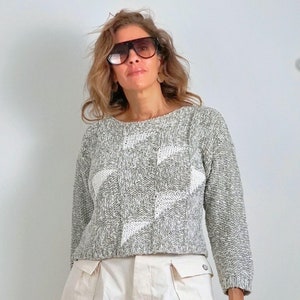 Puede incluir: Una mujer con un suéter de punto gris y blanco con un patrón geométrico y pantalones cortos color crema. El suéter tiene mangas largas y cuello redondo. Los pantalones cortos tienen bolsillos y cierre de botón. También lleva gafas de sol.