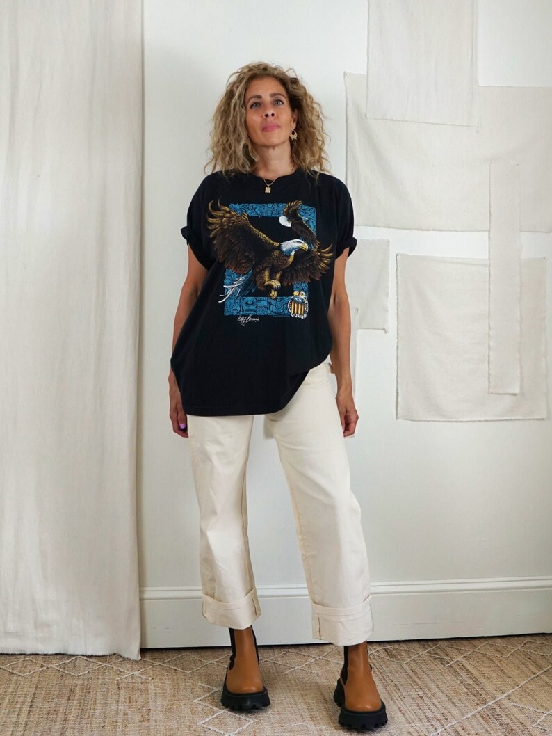 Op de afbeelding: Een zwart T-shirt met een afbeelding van een vliegende adelaar. De adelaar houdt een kleine Amerikaanse vlag in zijn klauwen. Het T-shirt wordt gecombineerd met een cr&egrave;mekleurige wijde broek en bruine enkellaarsjes.