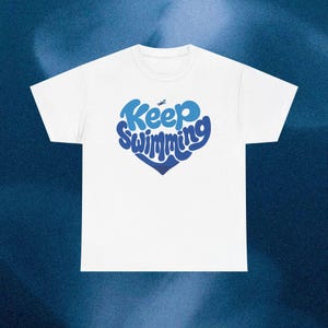 Könnte beinhalten: Weißes T-Shirt mit einem blauen herzförmigen Design mit der Aufschrift "Keep Swimming". Der Text ist in einer verspielten, stilisierten Schriftart gehalten. Über dem Wort "Keep" befindet sich eine kleine Schwimmer-Illustration.