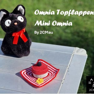 May include: A black plush cat with a red ribbon sits on a silver table next to a red and white crocheted hat with a red pom-pom. The text "Omnia Topflappe - Mini Omnia By 2CMau" is visible in the background.