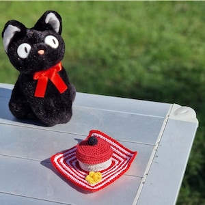 Op de afbeelding: Een zwarte pluche kat met een rode strik zit op een zilveren tafel naast een gehaakte rode en witte cupcake met een zwarte bes erop.