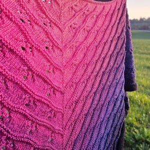 Peut inclure: Un châle tricoté à dégradé rose et violet. Le châle présente un motif texturé et ajouré avec des côtes diagonales. La couleur passe d'un rose vif à un violet profond, créant un accessoire visuellement attrayant.
