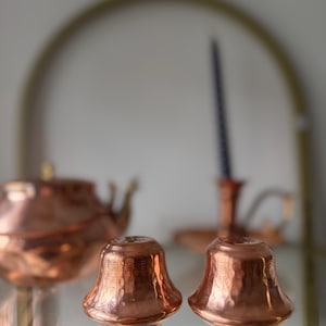 May include: Two copper-colored bell-shaped salt and pepper shakers with a hammered texture. A copper teapot and candle holder are visible in the background. The items are displayed on a glass shelf.
