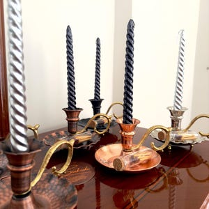 May include: A collection of decorative candle holders with spiral-shaped candles. The candle holders are made of copper and brass, with a vintage aesthetic. The candles are silver and black, adding contrast to the warm tones of the metal.