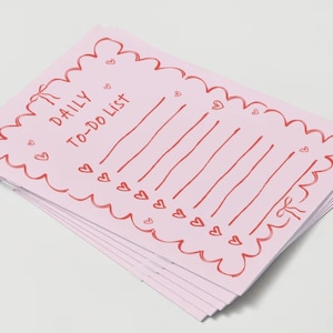 Puede incluir: Una pila de blocs de notas rosas para la lista de tareas pendientes con un borde rojo festoneado y corazones. El texto "DAILY TO-DO LIST" está escrito en rojo.