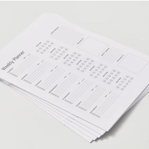 Puede incluir: Una pila de hojas de planificador semanal blancas. Cada hoja tiene secciones para cada día de la semana, con espacios para notas, seguimiento de la ingesta de agua y prioridades. El texto "Weekly Planner" está impreso en la parte superior.