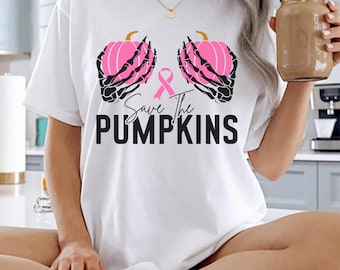 Op de afbeelding: Een wit T-shirt met korte mouwen en een afbeelding van twee zwarte skeletthanden die roze pompoenen vasthouden, met een roze lint in het midden. De tekst "Save The PUMPKINS" is in zwarte letters onder het ontwerp gedrukt.
