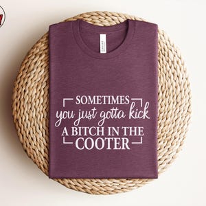 May include: A maroon t-shirt with the text "Sometimes you just gotta kick a bitch in the cooter" in white. The shirt is folded and placed on a woven, circular placemat. The brand logo is in the upper left corner.
