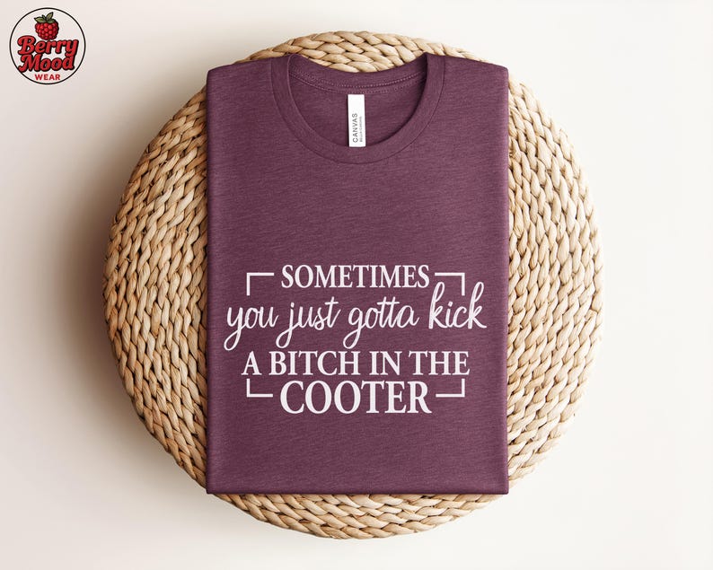 May include: A maroon t-shirt with the text "Sometimes you just gotta kick a bitch in the cooter" in white. The shirt is folded and placed on a woven, circular placemat. The brand logo is in the upper left corner.