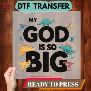 May include: DTF transfer with the text "MY GOD IS SO BIG" in large black letters, surrounded by colorful dinosaur illustrations. The words "READY TO PRESS" are at the bottom. The transfer is held against a wooden background.