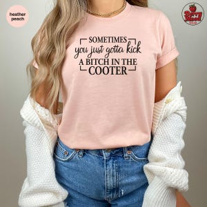 May include: Heather peach-colored t-shirt with the text "SOMETIMES you just gotta kick A BITCH IN THE COOTER." The shirt has rolled-up sleeves and is made of a soft material. The shirt is part of the Berry Mood Wear collection.