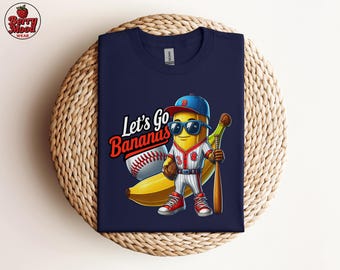 May include: Navy blue t-shirt featuring a cartoon banana dressed as a baseball player, wearing sunglasses and holding a bat. The shirt has the text "Let's Go Bananas" with a baseball graphic. The shirt is displayed on a woven surface.