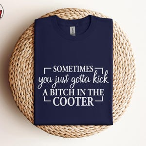 May include: Navy blue t-shirt with the text "SOMETIMES you just gotta kick A BITCH IN THE COOTER" in white. The shirt is displayed on a woven, circular surface. The Berry Mood Wear logo is in the upper left corner.