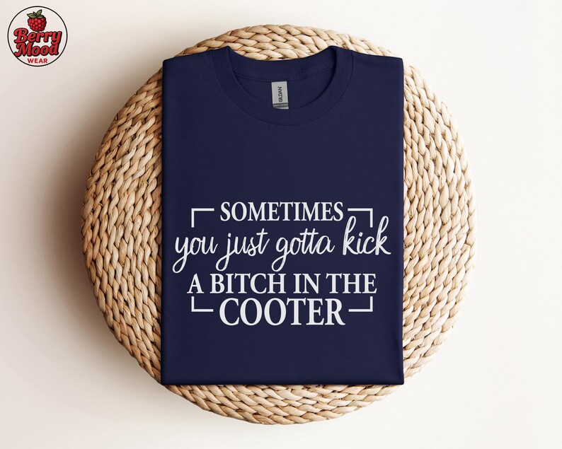 May include: Navy blue t-shirt with the text "SOMETIMES you just gotta kick A BITCH IN THE COOTER" in white. The shirt is displayed on a woven, circular surface. The Berry Mood Wear logo is in the upper left corner.