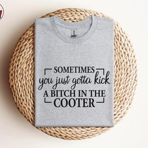 May include: A heather gray t-shirt with the text "SOMETIMES you just gotta kick A BITCH IN THE COOTER" in black. The shirt is displayed on a round, woven, light brown surface. The Berry Mood Wear logo is in the upper left corner.