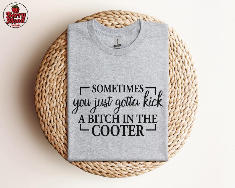 May include: A heather gray t-shirt with the text "SOMETIMES you just gotta kick A BITCH IN THE COOTER" in black. The shirt is displayed on a round, woven, light brown surface. The Berry Mood Wear logo is in the upper left corner.