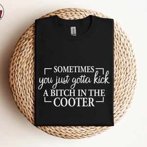 May include: Black t-shirt with the text "SOMETIMES you just gotta kick A BITCH IN THE COOTER" in white. The shirt is displayed on a round, woven, light brown surface. The Berry Mood Wear logo is in the upper left corner.