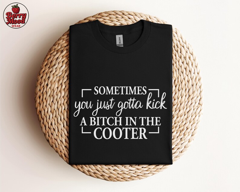 May include: Black t-shirt with the text "SOMETIMES you just gotta kick A BITCH IN THE COOTER" in white. The shirt is displayed on a round, woven, light brown surface. The Berry Mood Wear logo is in the upper left corner.