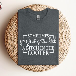 May include: Dark gray t-shirt with the text "SOMETIMES you just gotta kick A BITCH IN THE COOTER" in white. The shirt is displayed on a round, woven, light brown surface. The Berry Mood Wear logo is in the upper left corner.