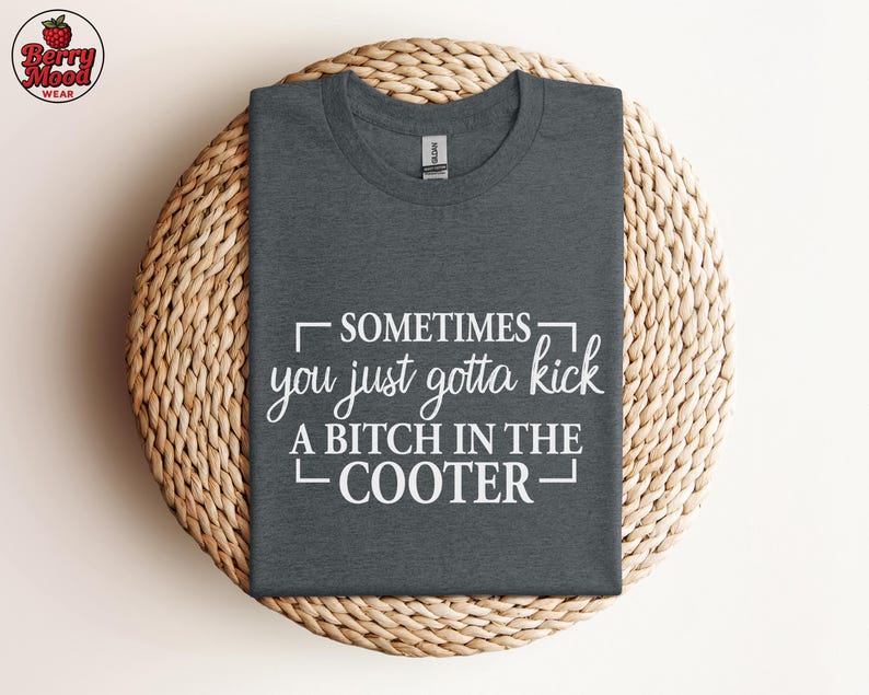 May include: Dark gray t-shirt with the text "SOMETIMES you just gotta kick A BITCH IN THE COOTER" in white. The shirt is displayed on a round, woven, light brown surface. The Berry Mood Wear logo is in the upper left corner.