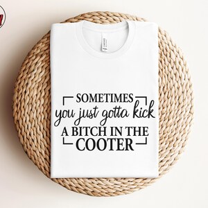May include: White t-shirt with the text "SOMETIMES you just gotta kick A BITCH IN THE COOTER" in black. The shirt is folded on a round, woven, light brown surface. The Berry Mood Wear logo is in the upper left corner.
