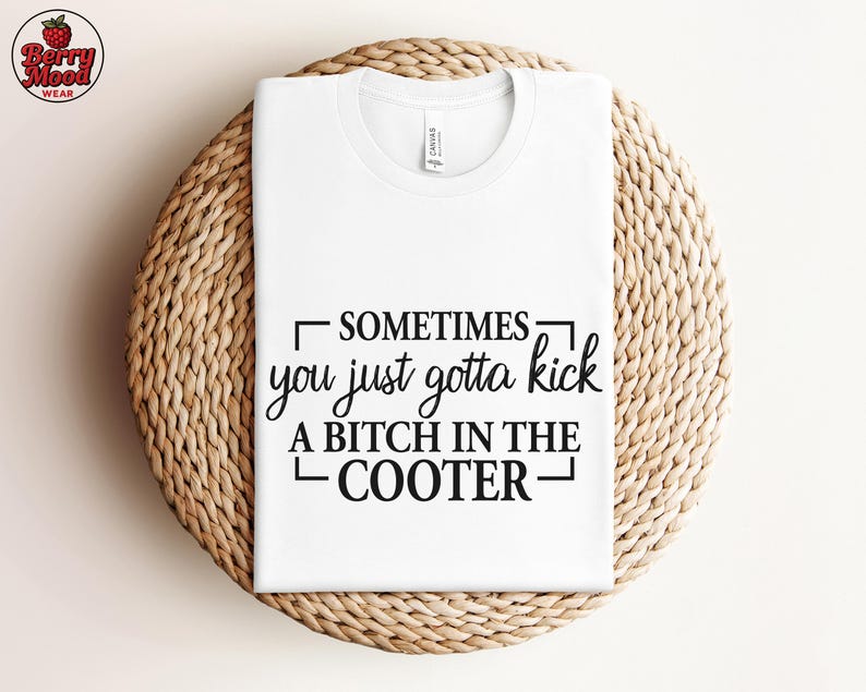 May include: White t-shirt with the text "SOMETIMES you just gotta kick A BITCH IN THE COOTER" in black. The shirt is folded on a round, woven, light brown surface. The Berry Mood Wear logo is in the upper left corner.