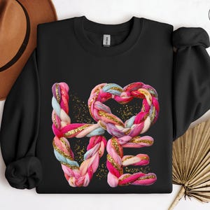 May include: Black crew neck sweatshirt with the word "LOVE" in a colorful, braided design. The "O" is a heart shape. Gold glitter accents the design. The sweatshirt has long sleeves and a ribbed neckline.