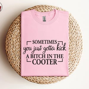 May include: A light pink t-shirt with the text "SOMETIMES you just gotta kick A BITCH IN THE COOTER" in black. The shirt is folded and placed on a woven, circular placemat. The Berry Mood Wear logo is in the upper left corner.