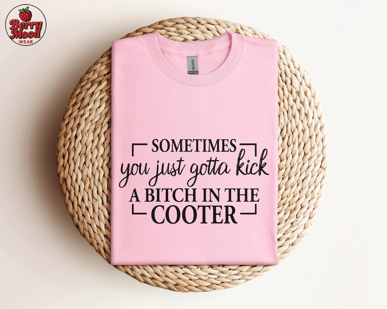 May include: A light pink t-shirt with the text "SOMETIMES you just gotta kick A BITCH IN THE COOTER" in black. The shirt is folded and placed on a woven, circular placemat. The Berry Mood Wear logo is in the upper left corner.