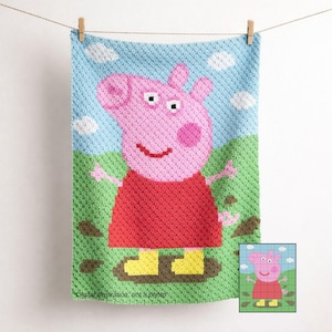 Peut inclure: Couverture crochetée représentant un cochon rose en robe rouge et bottes jaunes, debout dans la boue. Le fond comprend un ciel bleu avec des nuages blancs et de l'herbe verte. La couverture est suspendue à une corde à linge.
