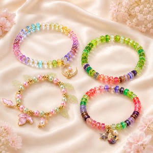 Può includere: Quattro braccialetti di perline con perle colorate, traslucide e opache. Un braccialetto ha un ciondolo a forma di luna dorata, uno ha un ciondolo a forma di stella e sirena dorata, e uno presenta perle rosa e verdi con foglie dorate.