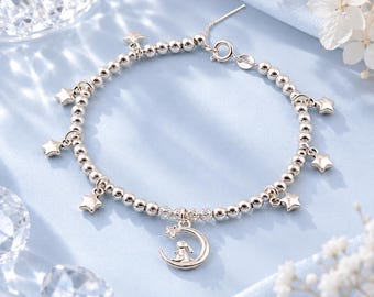 May include: A silver-toned beaded bracelet featuring multiple small star charms spaced along the chain. A central pendant depicts a small rabbit sitting inside a crescent moon shape. The bracelet includes a circular spring ring clasp closure.