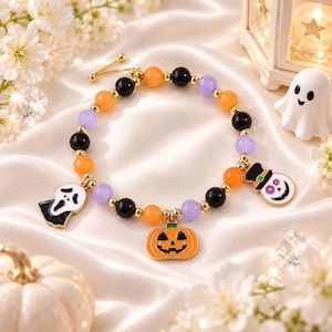 Peut inclure: Un bracelet composé de perles rondes alternant orange, noir et violet avec des intercalaires dorés. Le bracelet comporte trois breloques en émail : un masque de fantôme blanc, une citrouille d'Halloween orange et un crâne blanc avec un haut-de-forme.