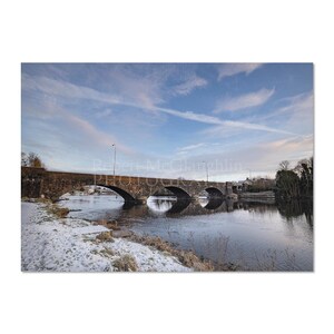 May include: A photograph of a stone bridge with multiple arches over a river. The sky is blue with streaks of cloud. Snow covers the riverbank. The bridge's reflection is visible in the water. The image has a tranquil feel.
