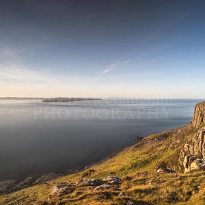 Può includere: Fotografia di un paesaggio di una scena costiera. L'immagine mostra una vasta distesa d'acqua sotto un cielo azzurro. Sulla destra una scogliera rocciosa con erba verde e rocce marroni. Un'isola lontana è visibile sullo sfondo.