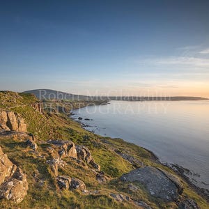 Può includere: Un paesaggio costiero panoramico con una scogliera rocciosa con vegetazione verde e grandi massi. Il mare calmo si estende fino all'orizzonte sotto un cielo blu con un bagliore dorato del tramonto. Fotografia di paesaggio.