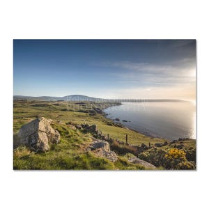 Può includere: Fotografia di un paesaggio costiero sotto un cielo azzurro. L'immagine presenta una costa rocciosa, colline verdi e il mare calmo. Il sole splende, creando un'atmosfera calda. Firmato Robert McCorquodale Photography.