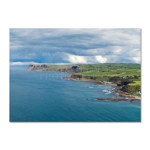 Può includere: Fotografia di un paesaggio costiero con un mare blu intenso, campi verdi e un cielo nuvoloso. L'immagine include la parola "PHOTO" al centro. Le scogliere e i campi sono visibili lungo la costa.