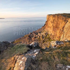 Può includere: Fotografia di un paesaggio con una scogliera rocciosa illuminata dalla calda luce del sole al tramonto. L'immagine cattura la vastità del mare e il cielo azzurro, con il testo "Robert McCaw PHOTO" visibile.