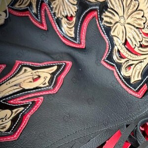 May include: Close-up of a black leather item with intricate floral designs in tan and red. The edges are trimmed with red stitching. The bottom features red leather fringe.