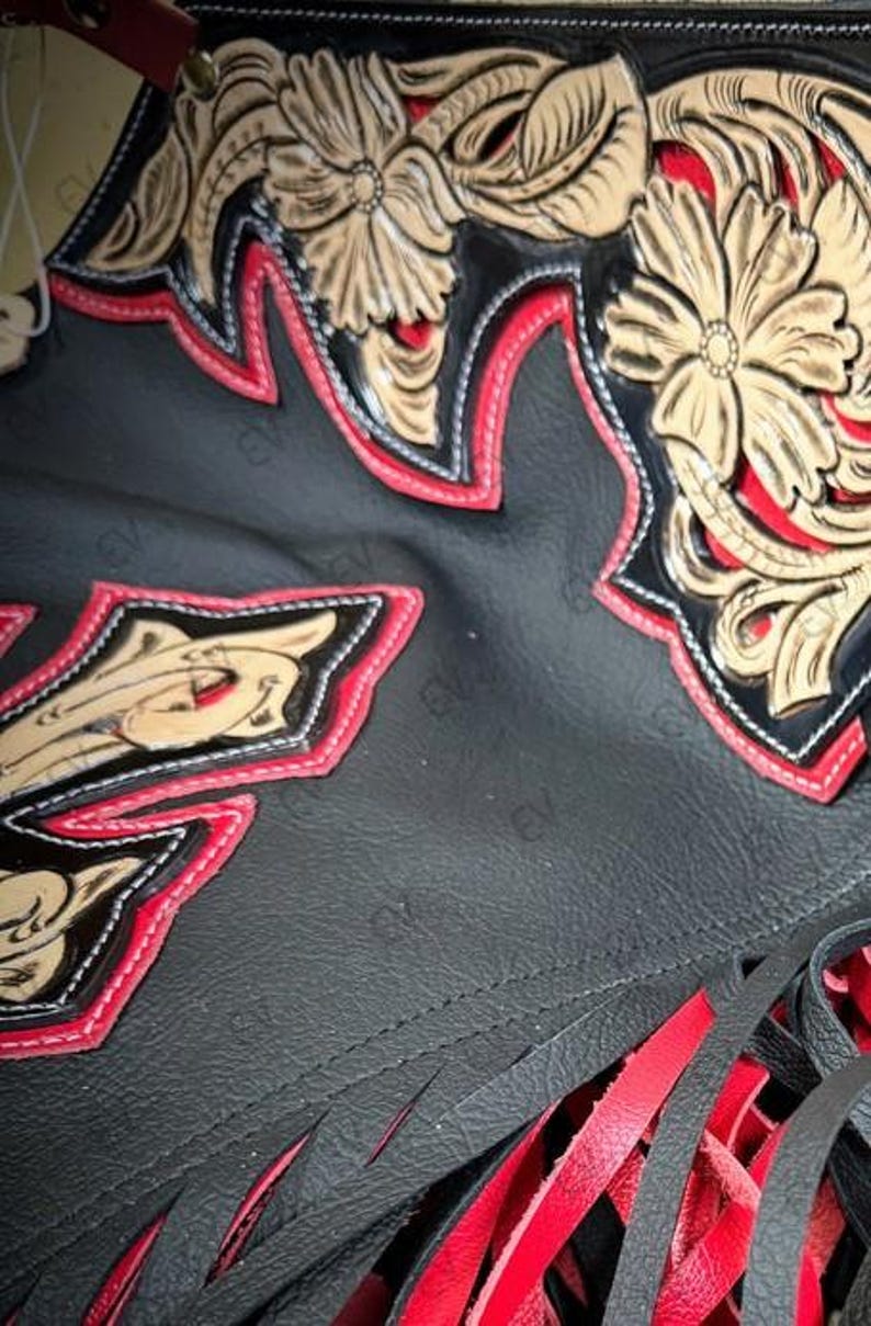 May include: Close-up of a black leather item with intricate floral designs in tan and red. The edges are trimmed with red stitching. The bottom features red leather fringe.