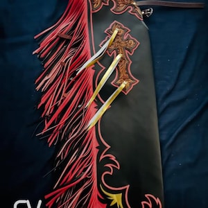 May include: Black leather chaps with red fringe and decorative cross and scrollwork. The chaps have a brown leather belt and are embellished with gold and white ribbons. The logo "EV Ravikings" is visible at the bottom.