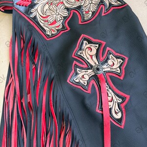 May include: Black leather chaps with red fringe and accents. Features include a decorative cross and floral design with red trim. The chaps have a leather strap and are designed for riding.