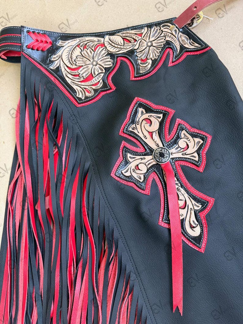 May include: Black leather chaps with red fringe and accents. Features include a decorative cross and floral design with red trim. The chaps have a leather strap and are designed for riding.